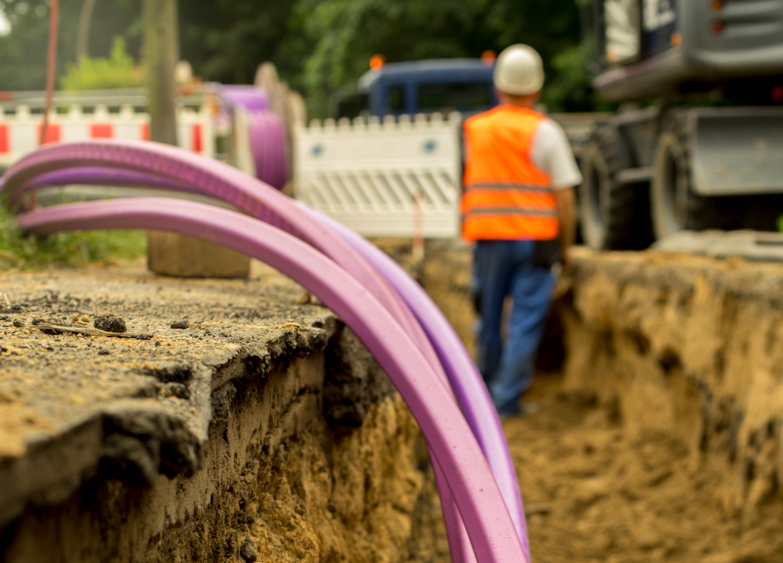 A construction worker in a safety vest and helmet stands near a deep trench with large purple pipes exposed, surrounded by construction equipment and barriers.
