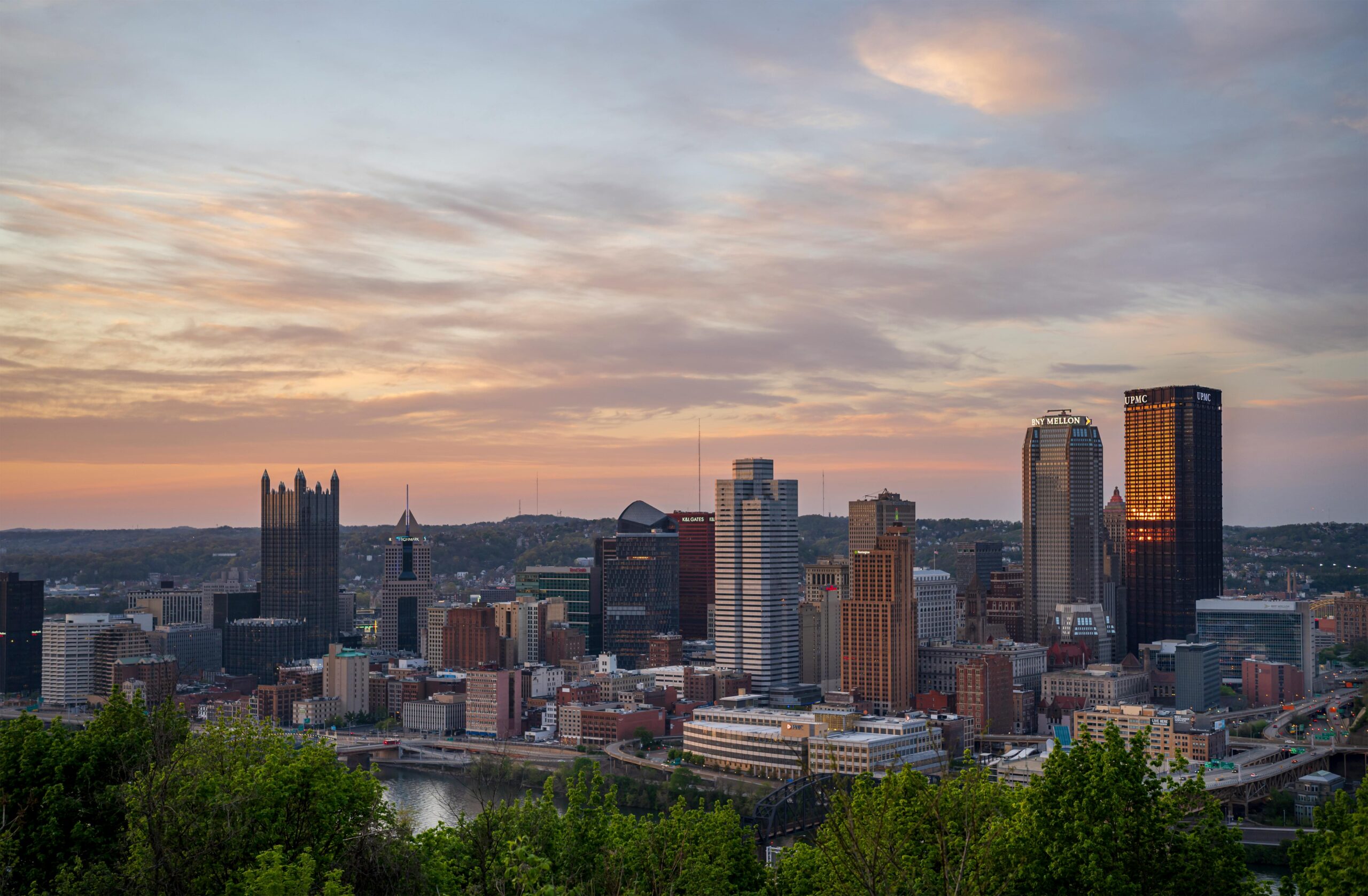 View of downtown Pittsburgh at sunset, featuring tall skyscrapers and historic buildings under a partly cloudy sky, with green trees and a river in the foreground—an ideal location for robust DAS solutions to enhance connectivity.