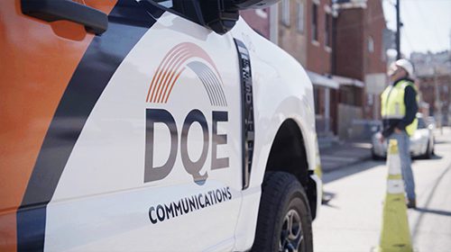 A DQE Communications work truck is parked on a city street. The truck’s door features the company logo. In the background, a worker in a safety vest and hard hat stands near traffic cones.