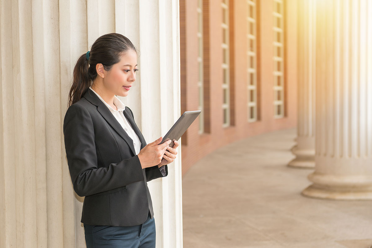 A woman in a black suit stands by large white columns, holding and looking at a tablet. The background shows a row of columns and windows, with warm sunlight streaming in.