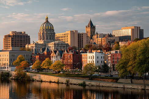 A cityscape featuring a domed capitol building, historic and modern structures, and autumn trees along a riverbank, all illuminated by warm, golden sunlight.