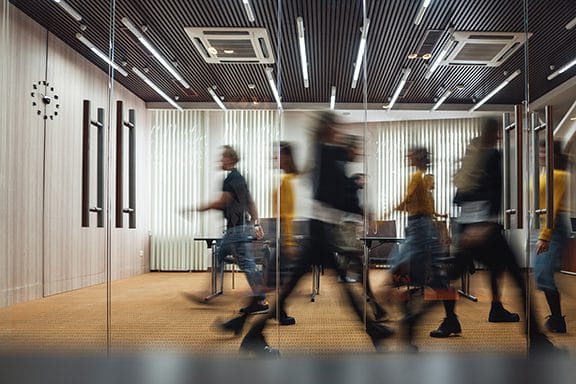Several people walk briskly through a modern office hallway with glass walls and a visible clock on the wall; their motion appears blurred.