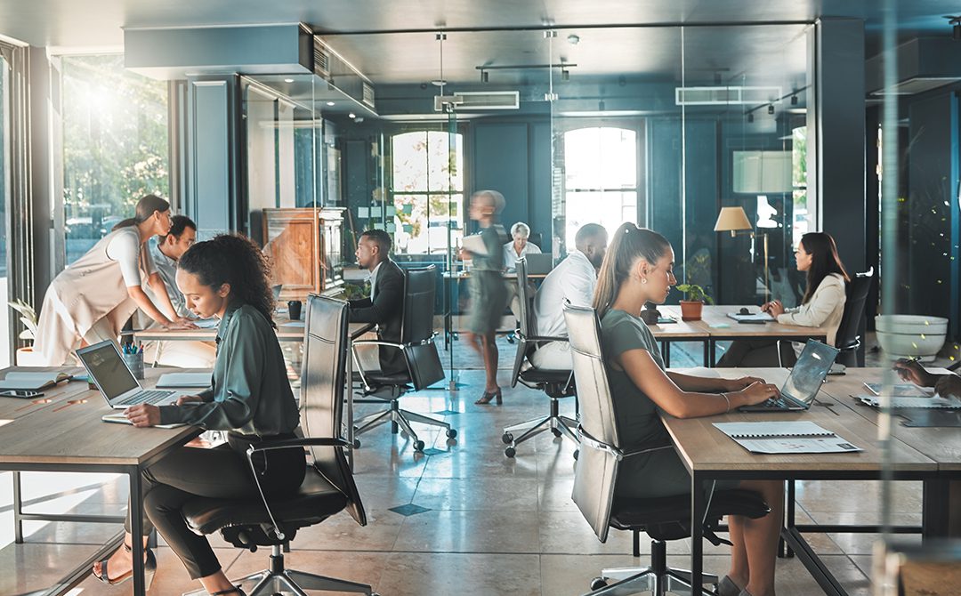 An open-plan modern office with several people working at desks on laptops and notebooks. Some are seated and focused on their work, while others are standing or walking, creating a collaborative, busy environment. Sunlight streams through large windows.