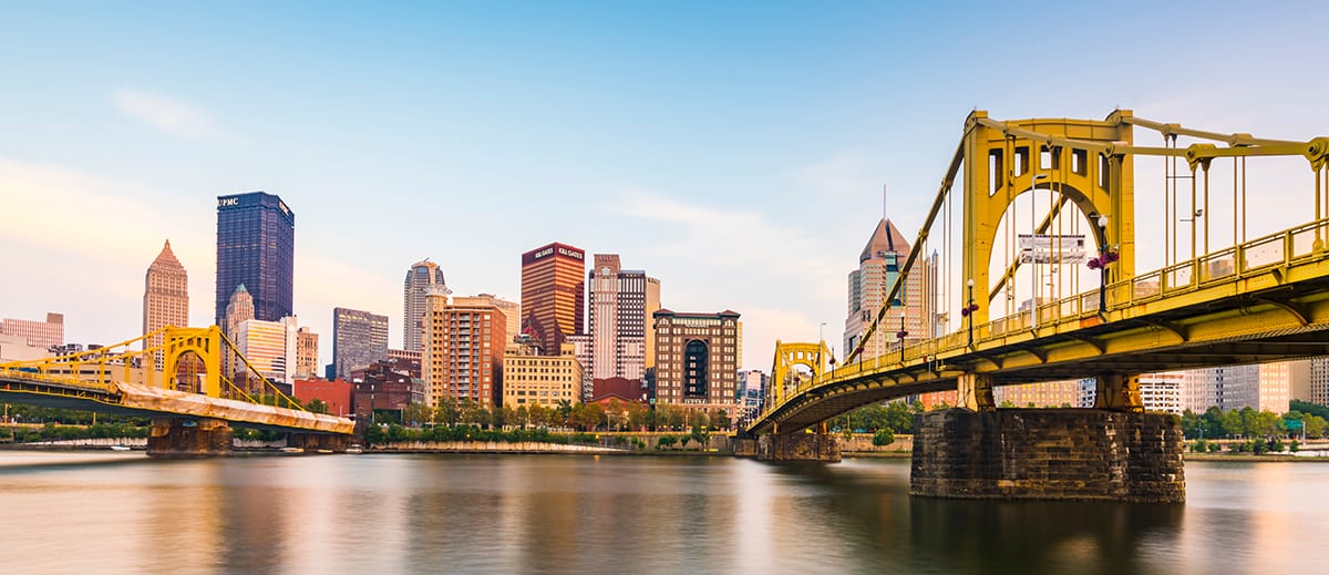 A yellow suspension bridge spans a calm river with a city skyline in the background under a clear sunset—symbolizing the reliable fiber connectivity in Altoona powered by DQE Communications.