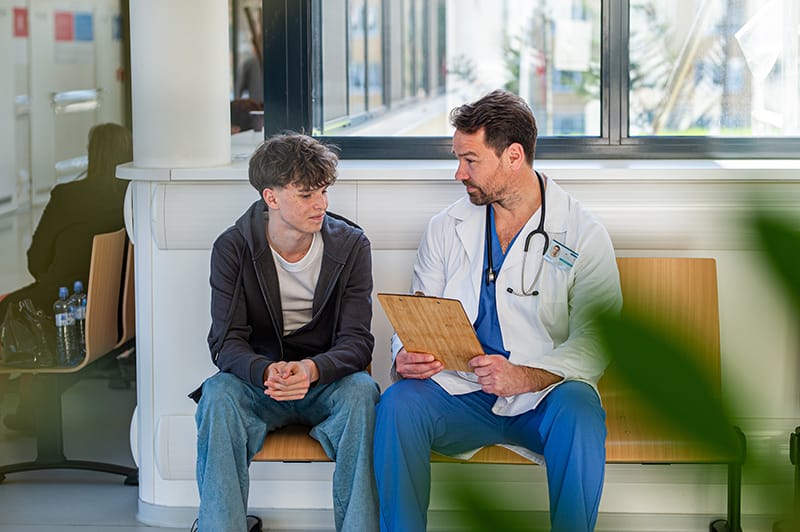 A doctor in a white coat and blue scrubs sits on a bench, holding a clipboard and talking with a young man in casual clothes. They are in a bright, modern hallway, part of an enterprise fiber Lancaster PA network, with sunlight streaming in.