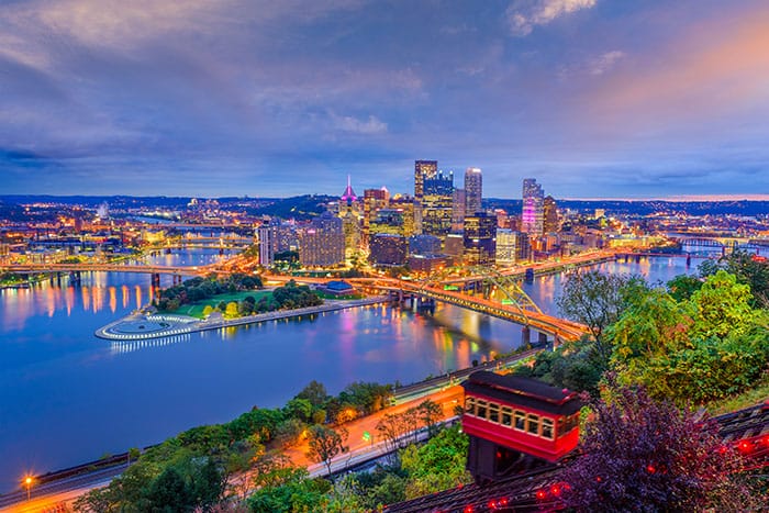 A vibrant Pittsburgh city skyline at dusk with illuminated buildings, bridges spanning rivers, and a red incline railway car in the foreground, surrounded by greenery and reflecting water—showcasing the spirit that drives DQE Communications to invest in Pittsburgh.